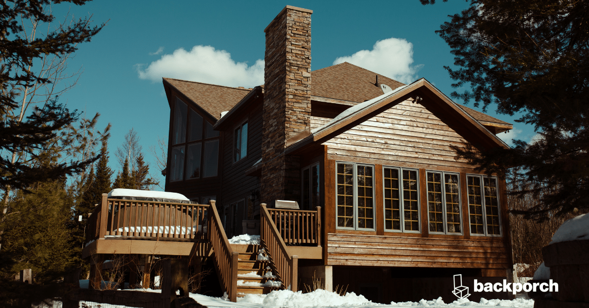 A cabin in the woods with a stone chimney and large deck is covered with a small amount of snow.