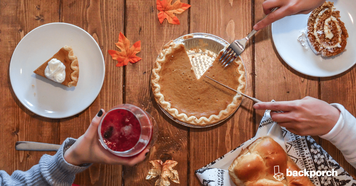 A Thanksgiving dinner guest holds up a cocktail near a loaf of bread while another guest slices a pumpkin pie.