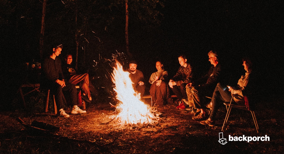 A group of seven adults are seated around a large bonfire in the woods.