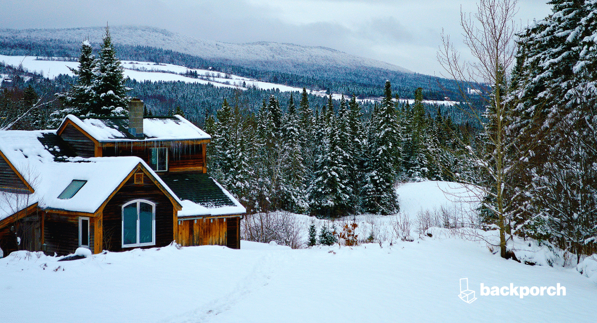 Snow covered landscape with a home surrounded by pine trees and a mountain range in the background.