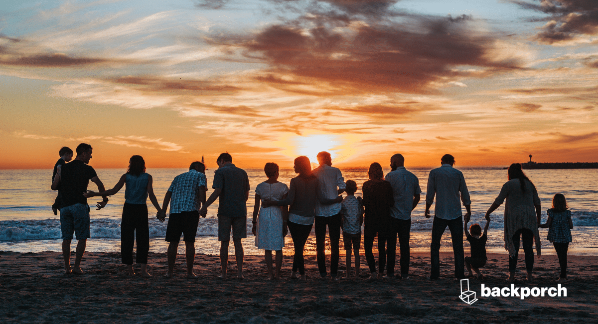 A large family of all ages gathered together on the shore of a beach facing the water at sunset.