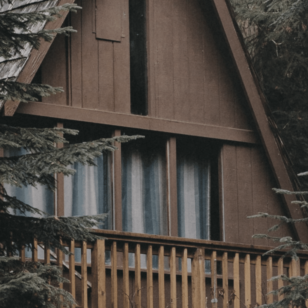A close-up of a wooden a-frame cabin with large windows and a balcony with a railing.