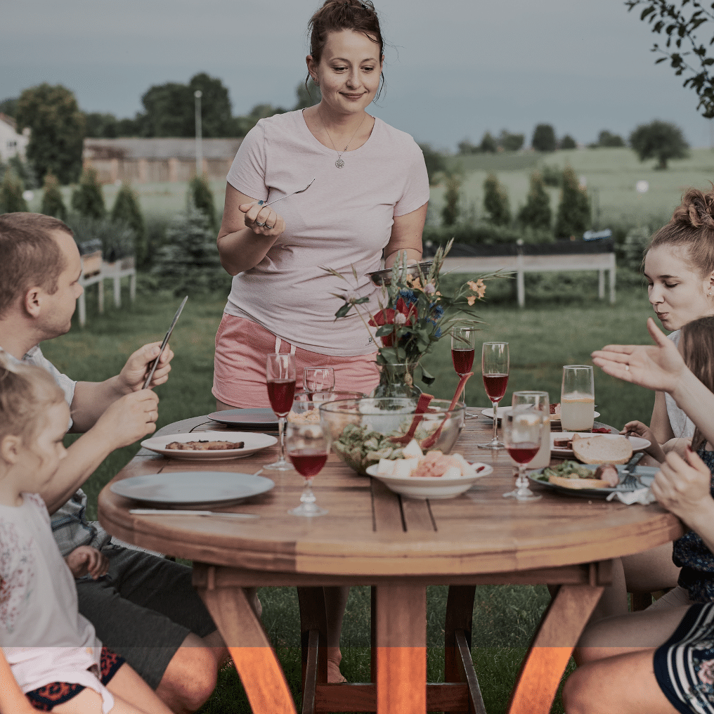 A woman standing at a picnic table with her family sitting around it eating a meal.