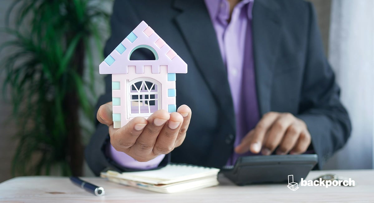 A man in a suit holding a model house while seated at a desk using a calculator.